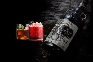 Botella de ron especiado y dos vasos de cócteles decorados con frutas y especias, sobre una superficie oscura con iluminación cálida y elegante.